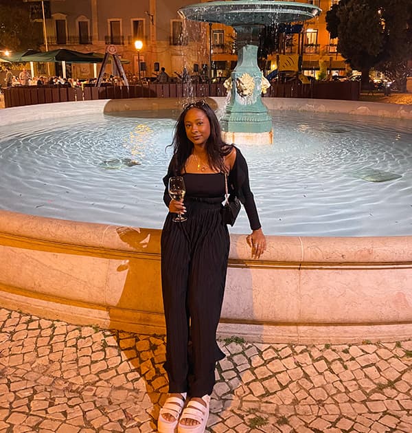 Jada Andrade standing in front of a lit fountain at night, holding a glass of wine and wearing a black outfit with white sandals.