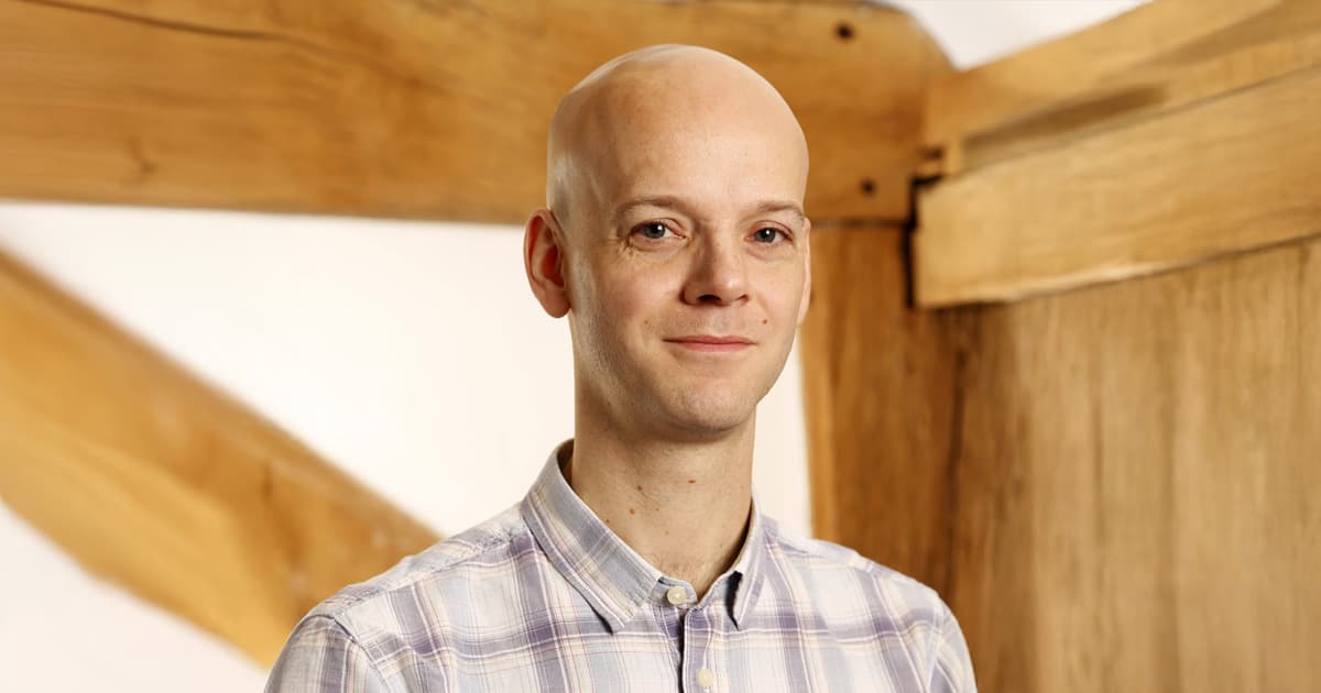 Jay Foad, wearing a light-coloured checked shirt, standing in front of a wooden beam structure, smiling softly at the camera