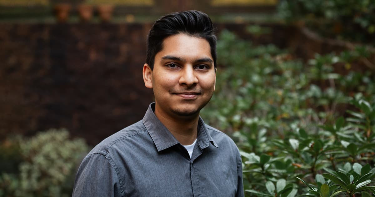 Josh David standing outdoors, smiling slightly, with greenery and a blurred stone wall in the background.