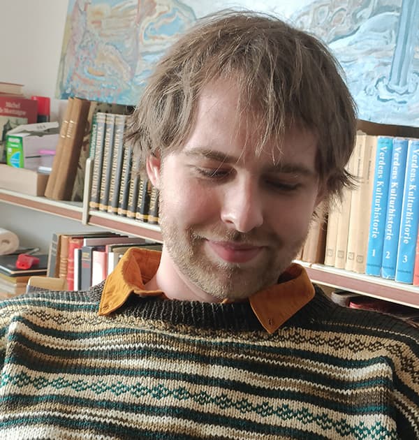 Karl Holt smiling and looking down, seated in front of a bookshelf filled with books and artwork.
