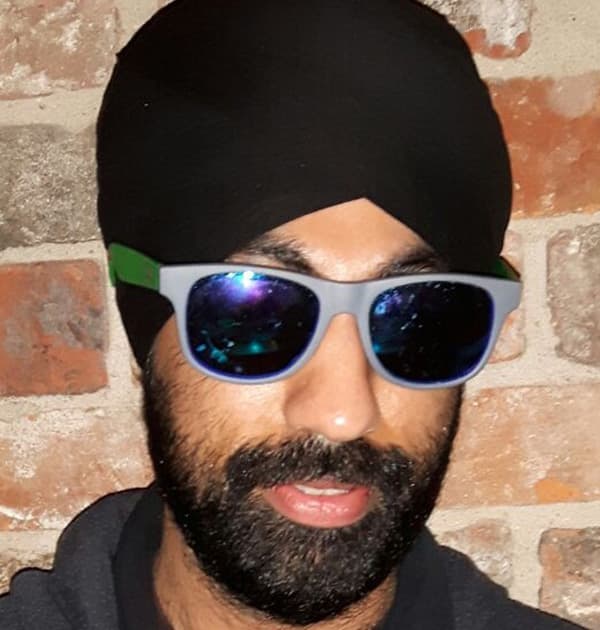 Karta Kooner wearing reflective sunglasses and a black turban, standing against a brick wall.