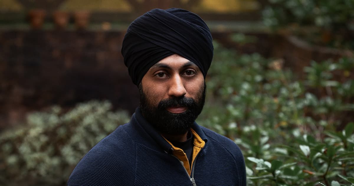Karta Kooner standing outdoors in front of greenery, wearing a dark turban and navy zip-up top.