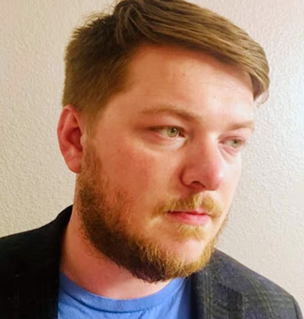 Nathan Rogers looking to the side, wearing a blue shirt and blazer against a neutral wall.