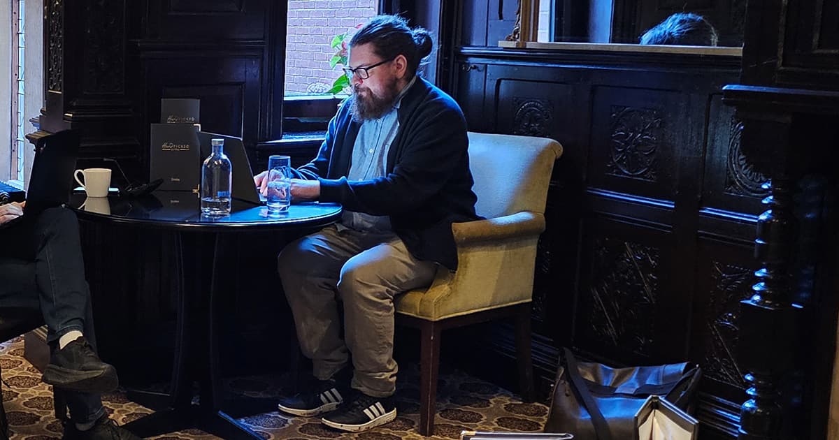 Neil Kirsopp sitting at a round table with a laptop and glass of water in a dark wood-panelled room.