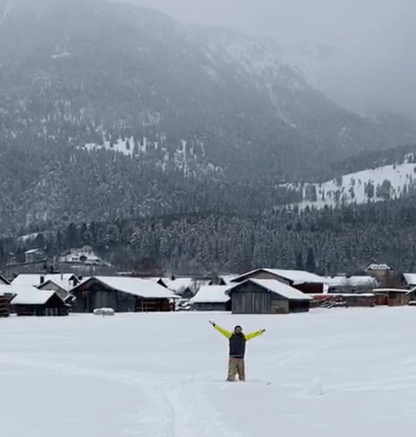 Neil Kirsopp standing with arms raised in a snow-covered field with alpine cabins and forested mountains in the background.