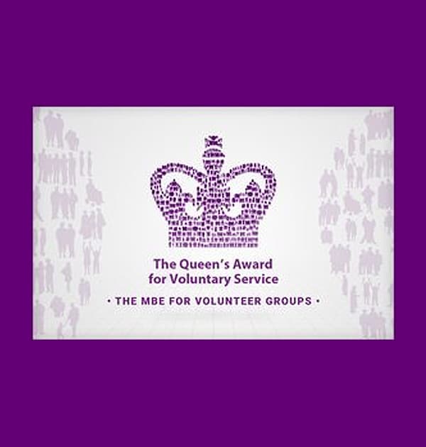 The Queen’s Award for Voluntary Service logo featuring a purple crown made of small human figures, with the text “The MBE for Volunteer Groups.”