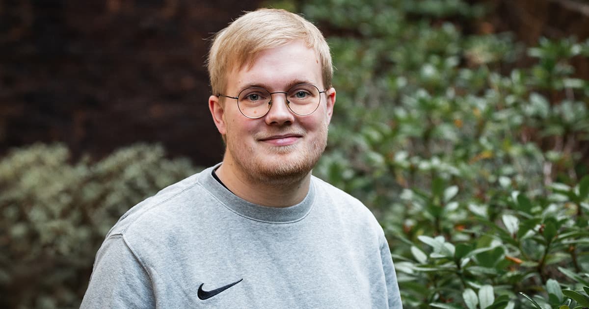 Peter Mikkelsen standing outdoors in front of green foliage, wearing a grey Nike sweatshirt and glasses.