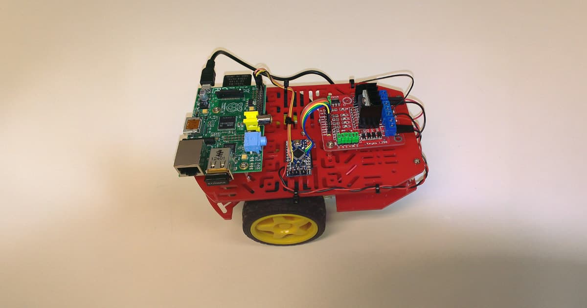 A small robot built with a red chassis, equipped with a Raspberry Pi, circuit boards, and various electronic components, sitting on a white surface.