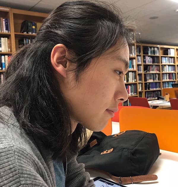 Shuhao Yang working on a tablet in a library, surrounded by bookshelves and orange chairs.