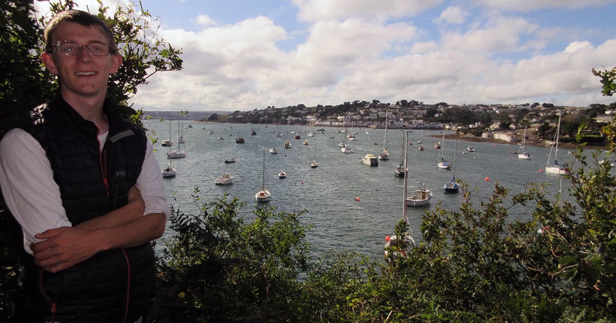 Silas Poulson smiling with arms crossed in front of a scenic harbour filled with boats, with a town visible across the water.