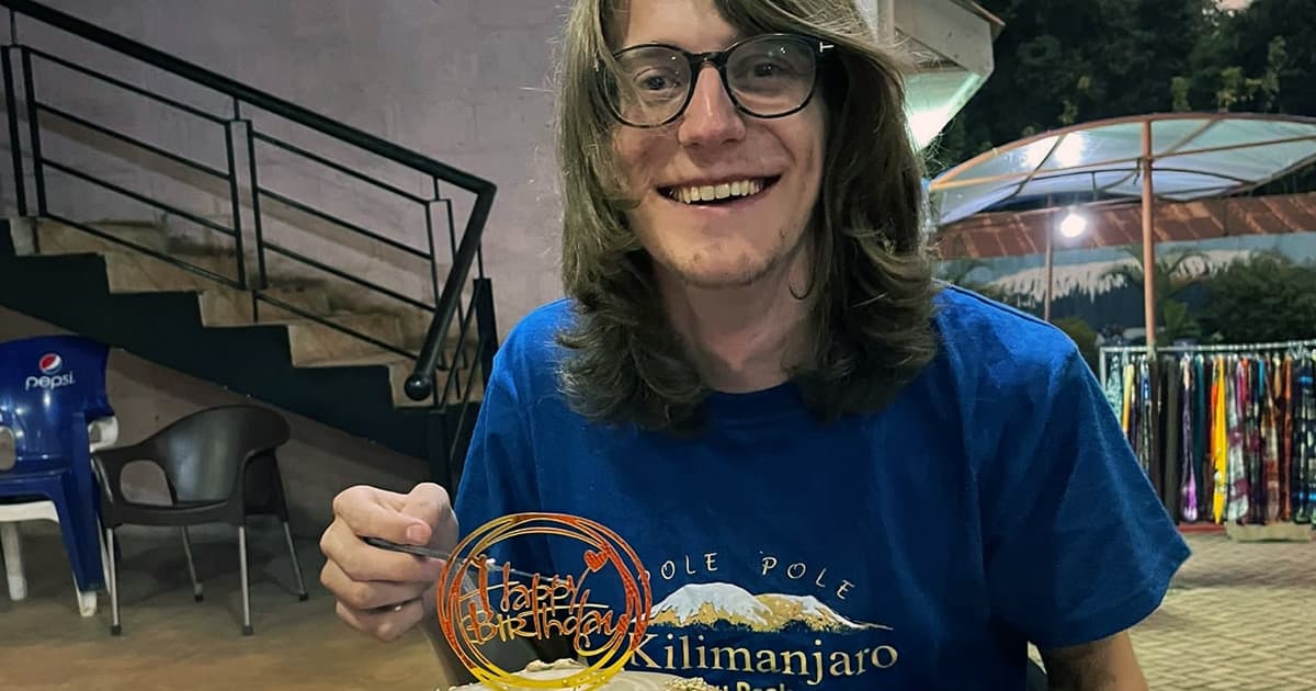 Silas Poulson smiling and holding a dessert with a “Happy Birthday” sign, wearing a Kilimanjaro t-shirt at an outdoor venue.