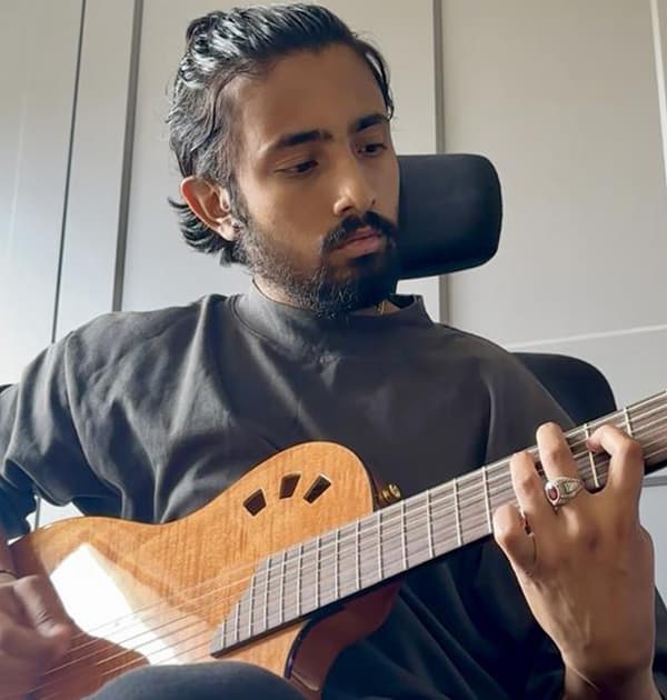Abs Suri sitting in a chair, focused while playing a wooden electric-acoustic guitar in a room with white wardrobes behind him.
