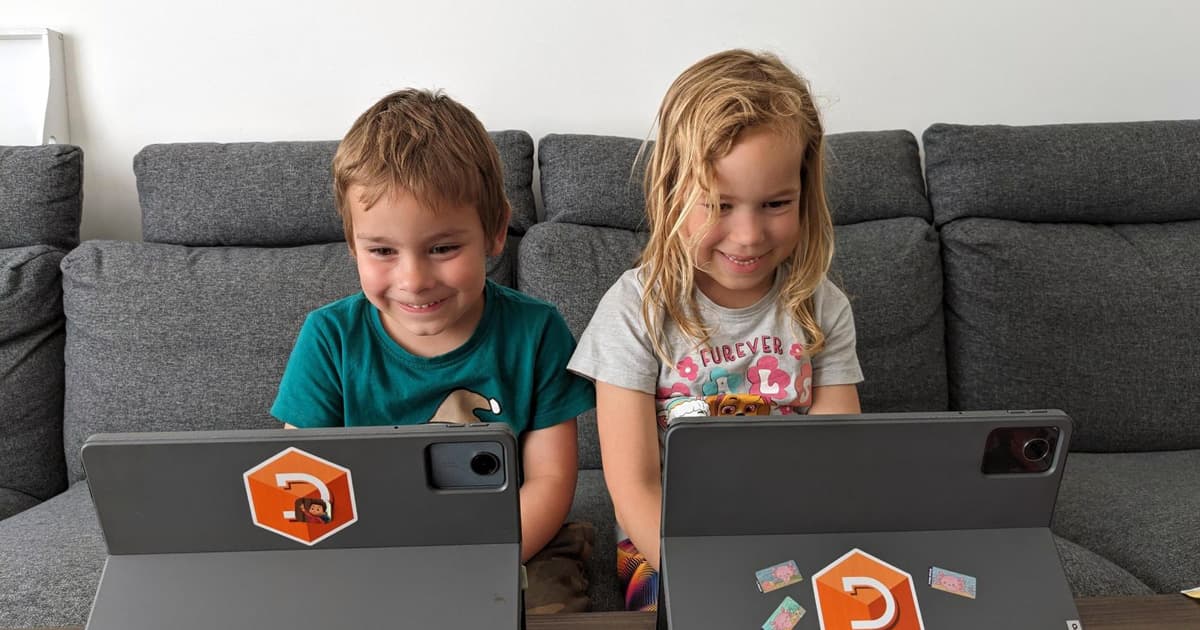 Two young children smiling and using tablets on a grey sofa, each device decorated with a Dyalog APL sticker and other colourful stickers.