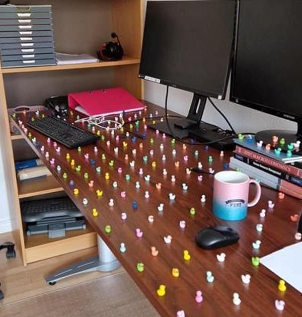 Office desk covered with hundreds of tiny colourful rubber ducks arranged in a grid pattern, surrounded by computer monitors, a keyboard, stationery, and a mug.