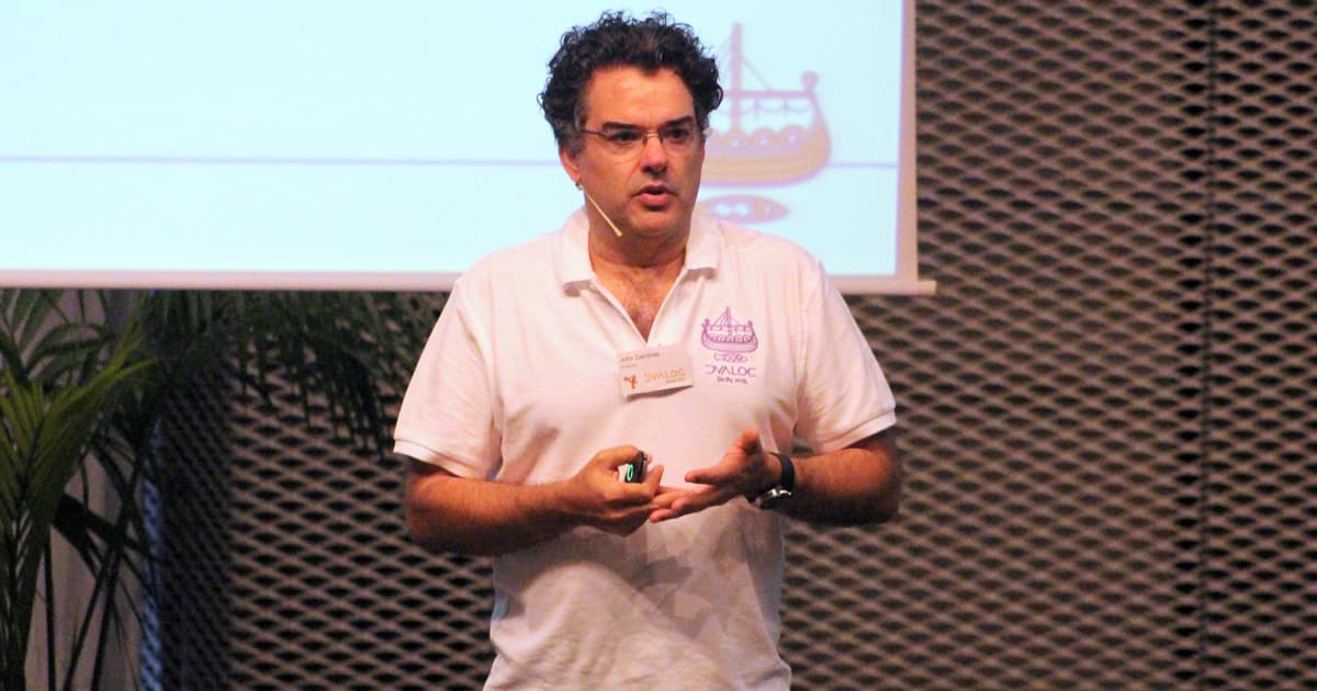John Daintree, wearing a white Dyalog ’15 polo shirt, speaking during his presentation in Sicily with a projected slide behind him.