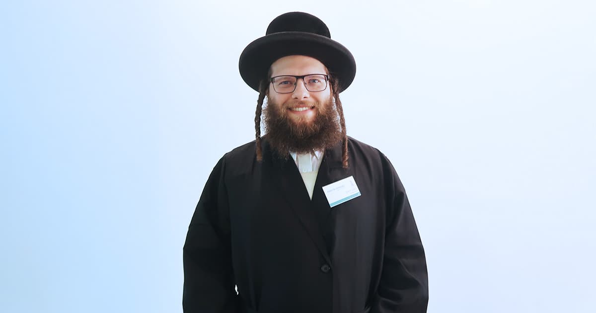 Adám Brudzewsky, smiling and wearing traditional attire, including a black coat and hat, with glasses and a name tag, standing against a light blue background.