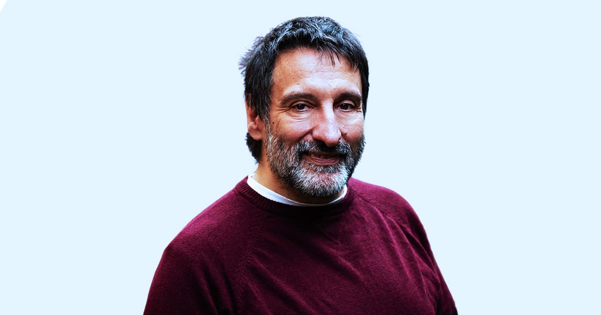 Michael Baas, smiling and wearing a maroon jumper over a white shirt, standing against a light blue background.