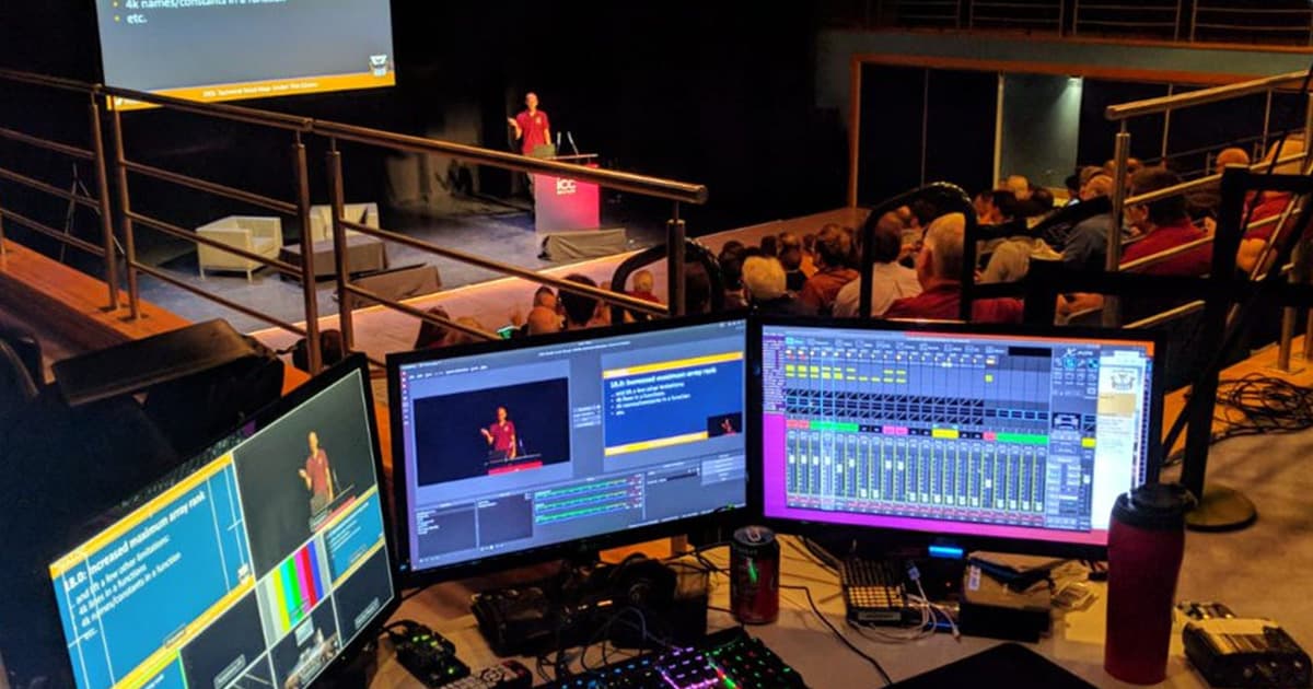 Control desk view at Dyalog ’18 showing multiple screens managing video, slides, and audio while a presenter speaks on stage to a full audience.