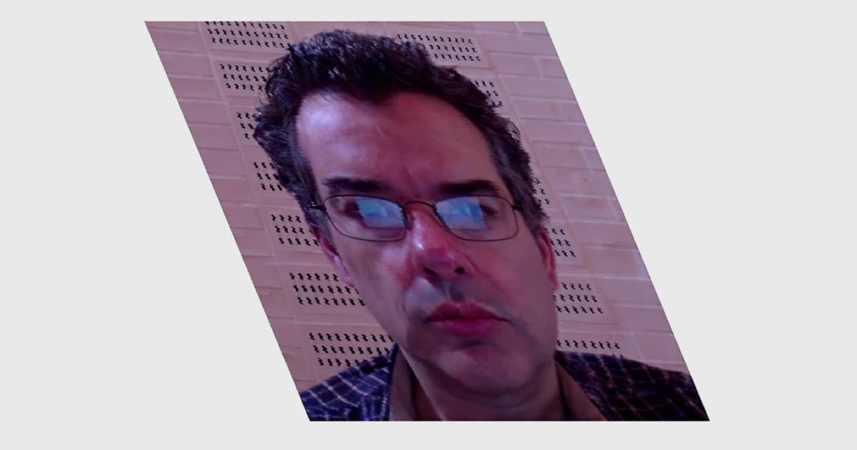 A tilted image of John Daintree at Dyalog ’14, with a geometric distortion effect applied. He is wearing glasses and a checked shirt against a perforated wall background.