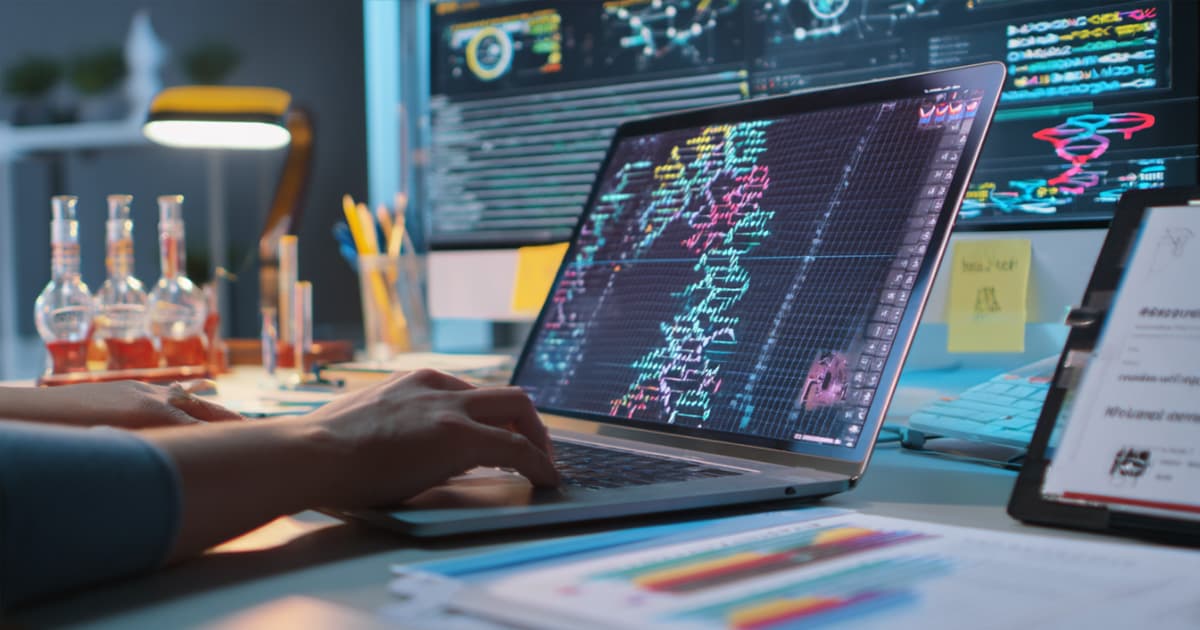 Close-up of a person working on a laptop displaying colourful DNA sequence data in a bioinformatics application, with scientific equipment, documents, and multiple monitors showing complex visual data in the background.