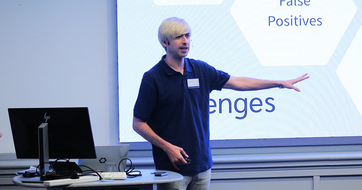 Brandon Wilson presenting at Dyalog ’24 in Glasgow. He stands in front of a projected slide with the word “Challenges,” wearing a dark polo shirt, gesturing with his right arm extended while speaking.
