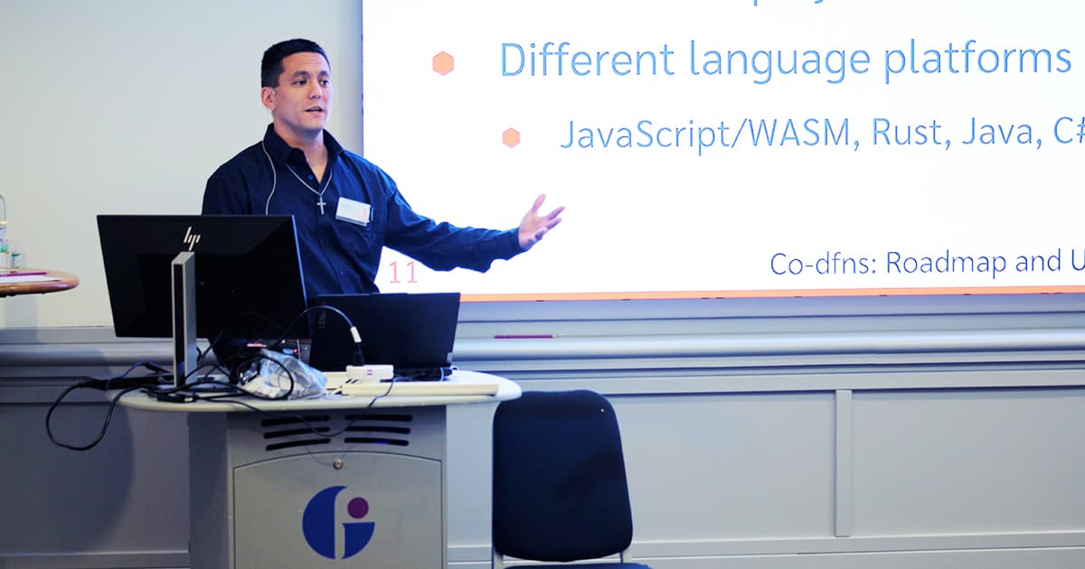 Aaron Hsu presenting at Dyalog ’24 in Glasgow. He stands at the podium in front of a slide listing “Different language platforms: JavaScript/WASM, Rust, Java, C#.” He gestures with his right hand while explaining the Co-dfns roadmap and updates.