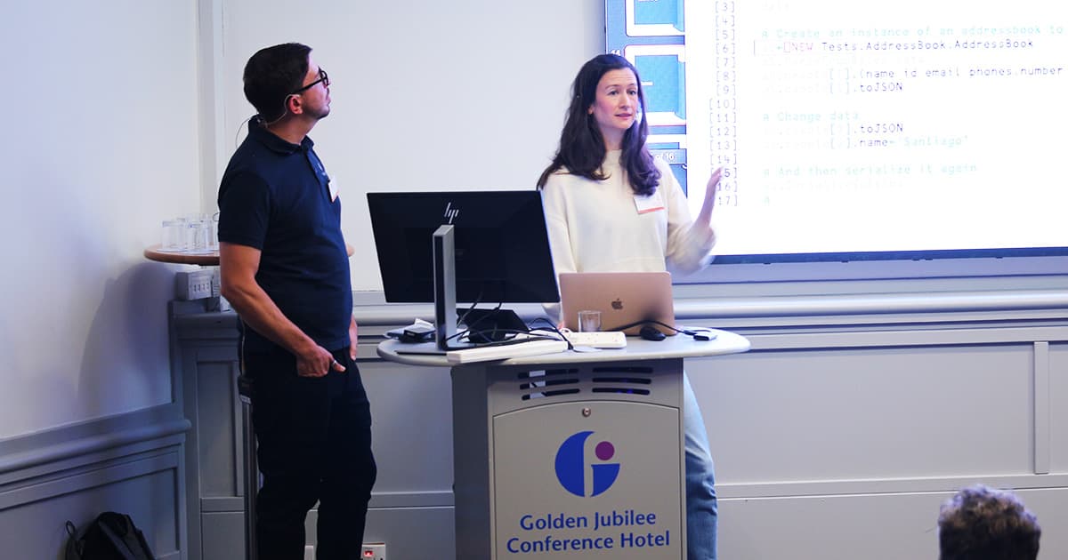 Gilgamesh Athoraya and Sandra Persson presenting together at Dyalog ’24 in Glasgow. Sandra stands at the podium, speaking with hand raised, while Gilgamesh looks on. The projected screen shows APL code demonstrating serialization. A Golden Jubilee Conference Hotel lectern is in front of them.