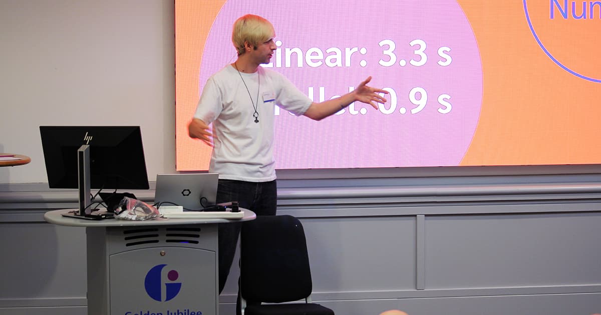 Brandon Wilson presenting at Dyalog ’24 in Glasgow. He gestures towards a bright slide behind him showing performance timings (“Linear: 3.3s”, “Mult: 0.9s”). A laptop and monitor sit on the lectern with cables and devices connected.