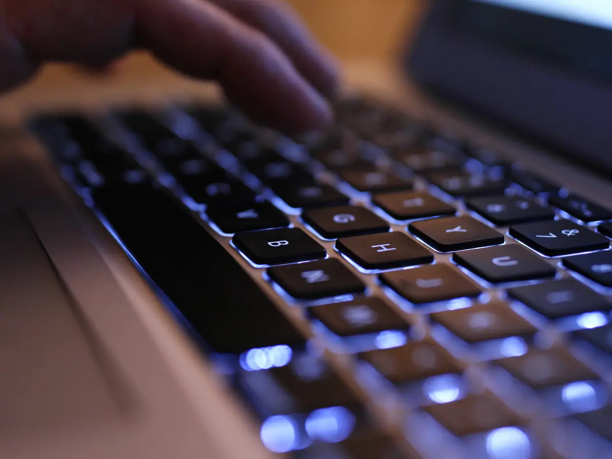 Close-up of a person’s hand typing on a laptop keyboard with blue backlit keys, photographed at a shallow angle with soft focus and warm ambient lighting.