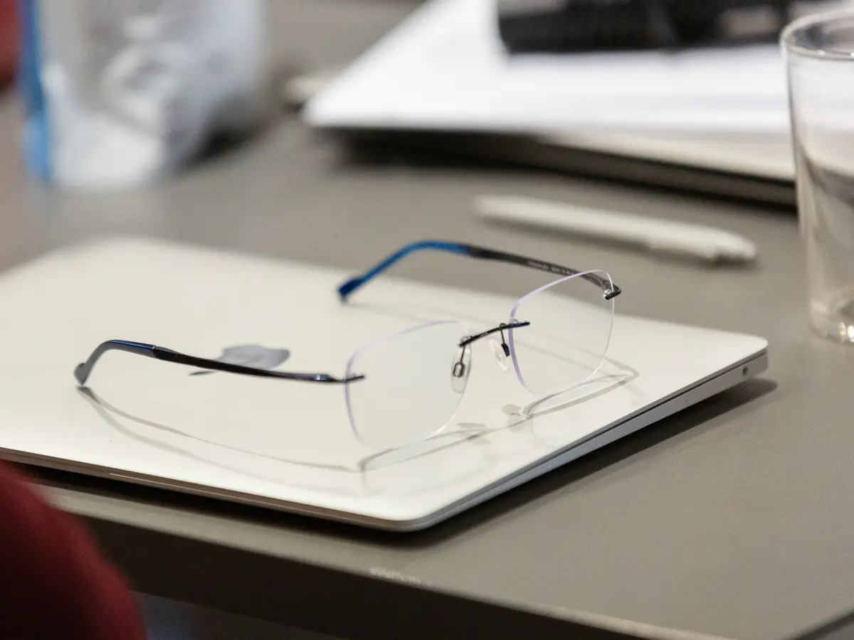Pair of rimless glasses resting on a closed silver laptop on a desk, with a glass of water and blurred office items in the background.