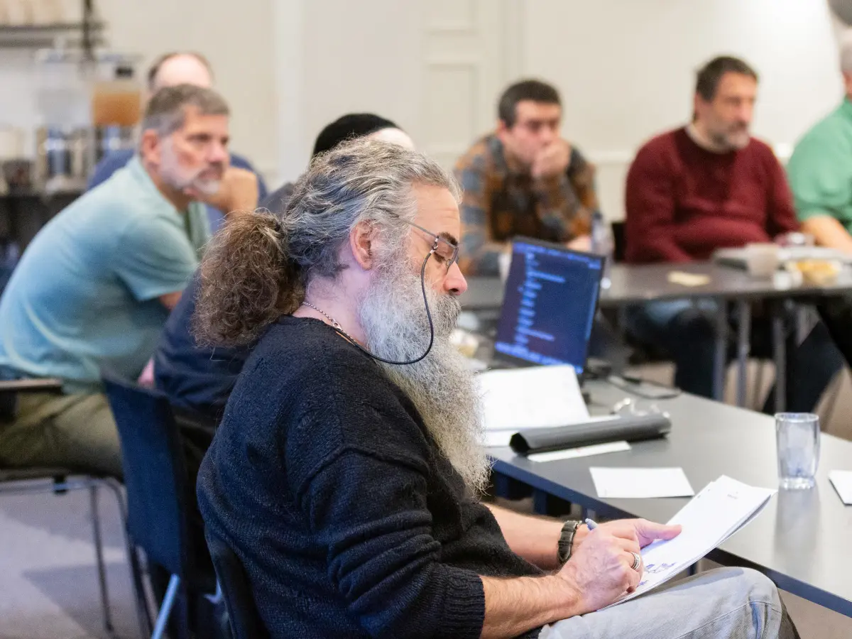John Daintree, with long grey hair and a beard, taking notes in a notebook while seated at a table during a group workshop, with other attendees and laptops visible in the background.