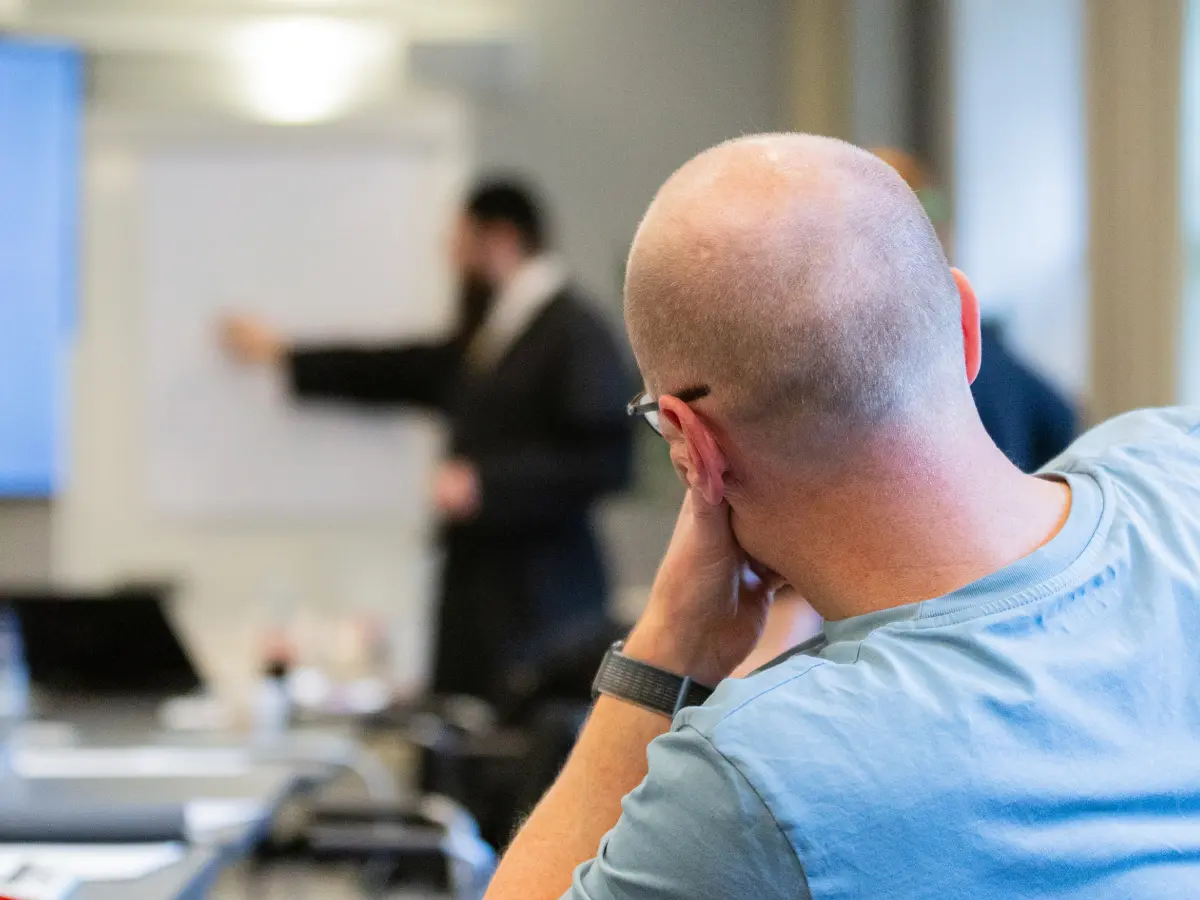 Rear view of an attendee seated at a table, resting their head on one hand while watching a presenter gesturing toward a whiteboard at the front of the room.