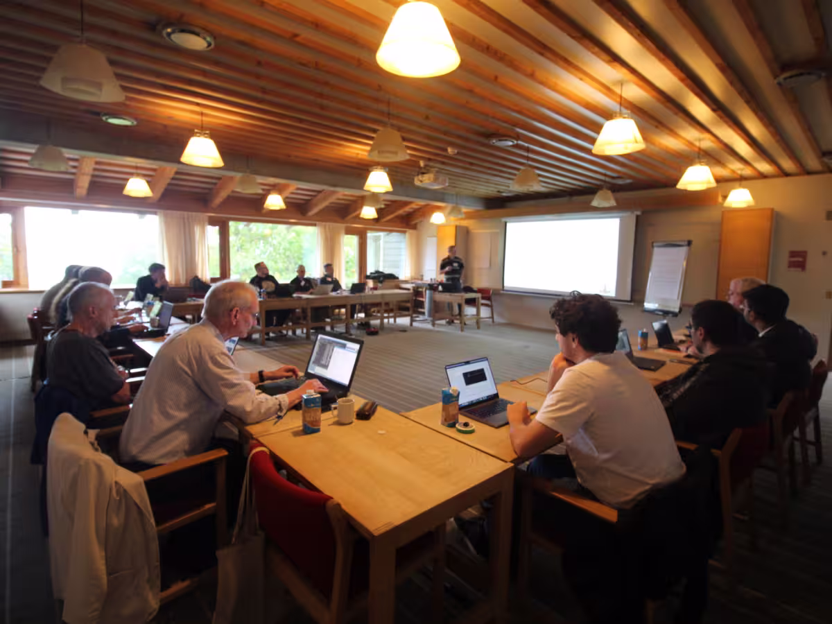 Attendees taking part in a Dyalog workshop, seated around tables with laptops in a large room with a projector screen at the front.