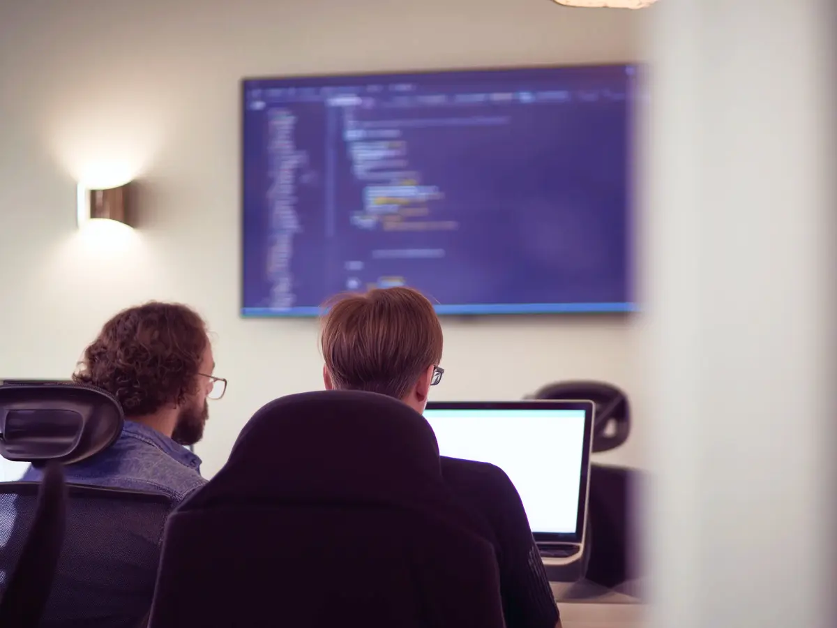 Two people seated at desks working on computers in an office environment, with a large screen in the background displaying blurred source code or technical content.
