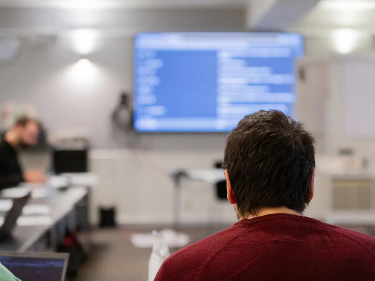 A person seen from behind sitting in a training or meeting room, facing a large screen displaying blurred code or technical content, with other attendees working at desks in the background.