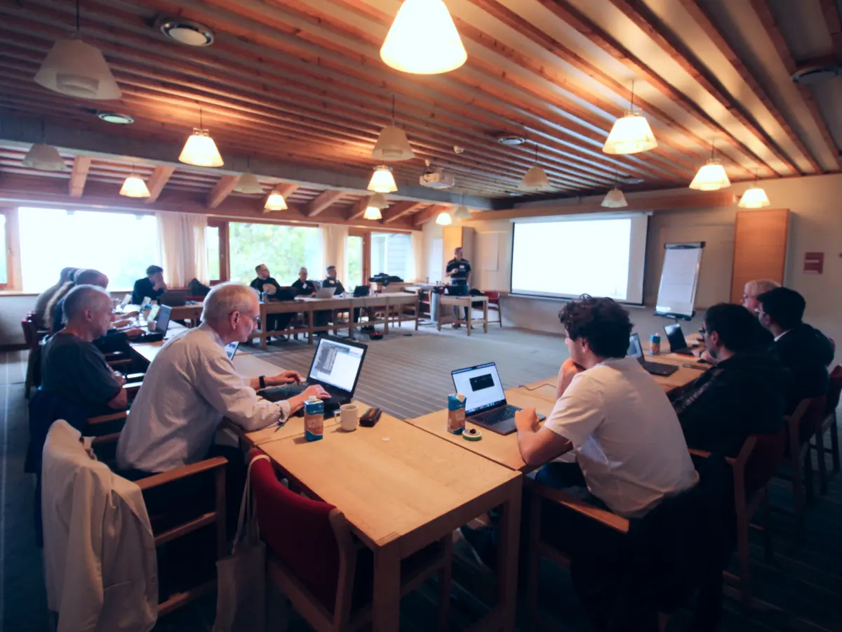 A group of people seated around tables in a large meeting room, working on laptops during a training session while a presenter stands at the front beside a projected screen.