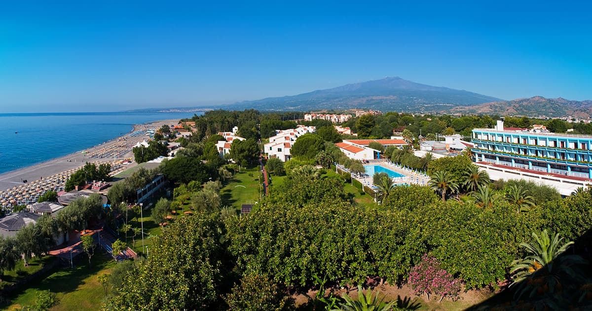 Aerial view of Atahotel Naxos Beach in Giardini Naxos, showing the resort's buildings, swimming pool, gardens, beach with sun loungers, and Mount Etna in the background.
