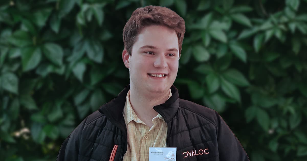 Hoover smiling, wearing a black Dyalog jacket and a light-coloured checked shirt, with a name badge visible, standing in front of a leafy green background.