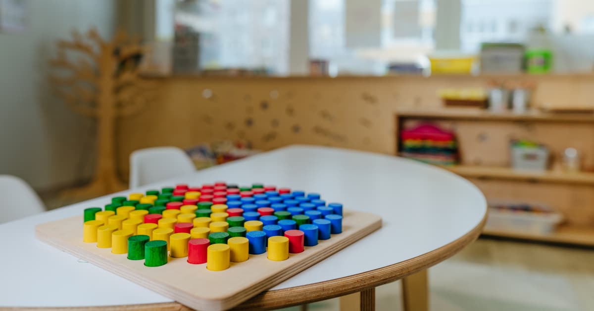 A wooden board game with rows of colourful round pegs in red, yellow, green, and blue, placed on a white table in a classroom setting.